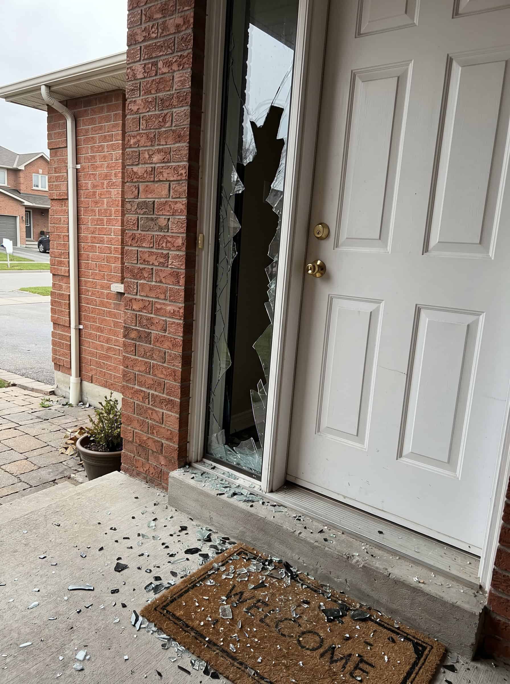 Real job photo: a front entry sidelite smashed out beside a white front door, shards across the porch and the welcome mat