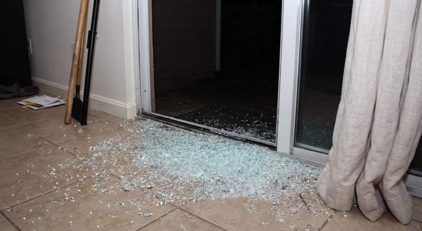 Real job photo: broken glass spilled across interior tile inside a patio door after a break-in, a broom resting against the wall