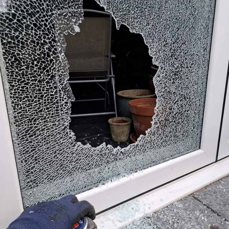 A sliding patio door with a ragged hole punched clean through the tempered glass during a break-in, garden chairs and pots visible inside and a gloved technician hand at the sill